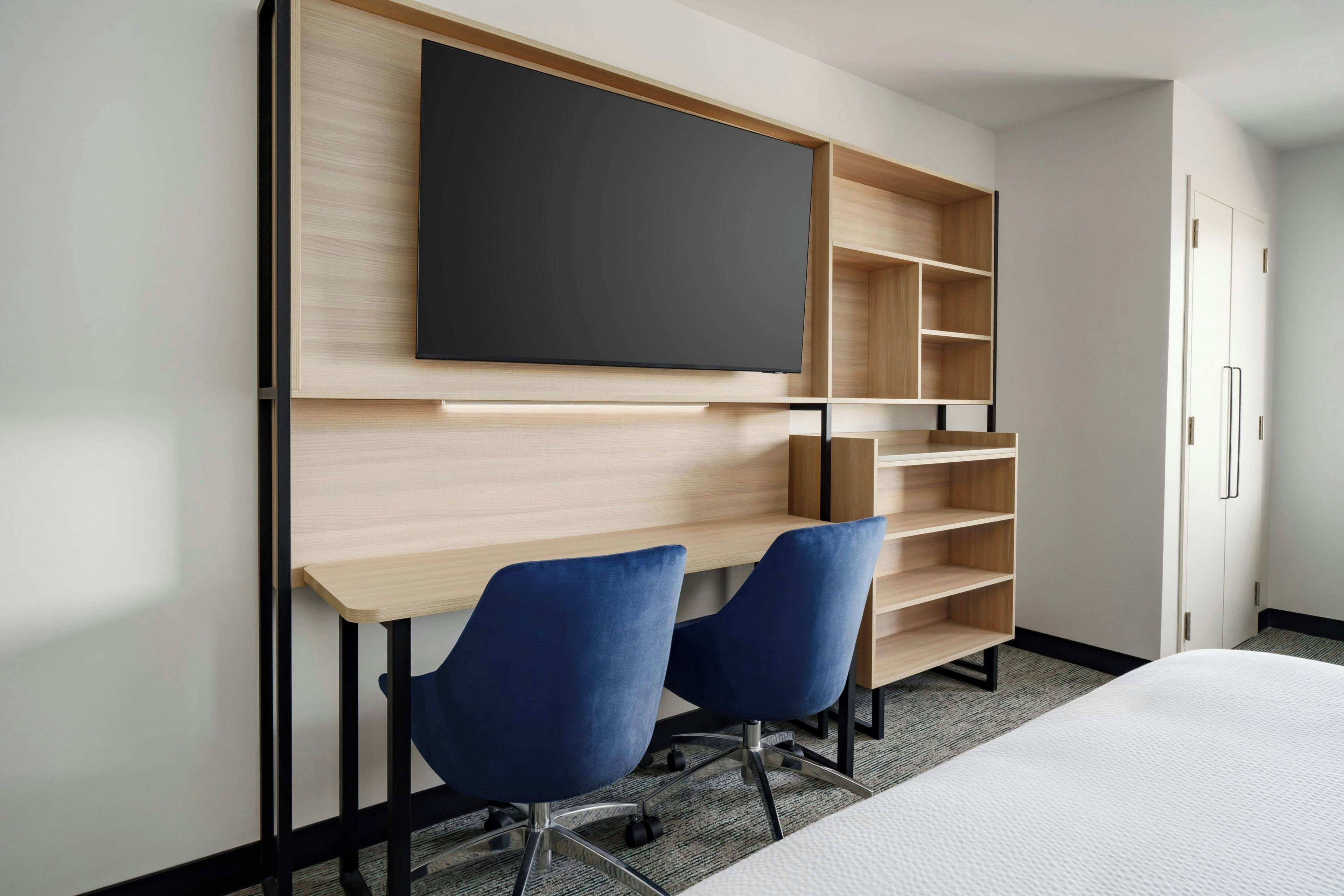 Quarto de hotel com cama, escrivaninha e duas cadeiras, televisão de tela plana na parede e prateleiras embutidas.