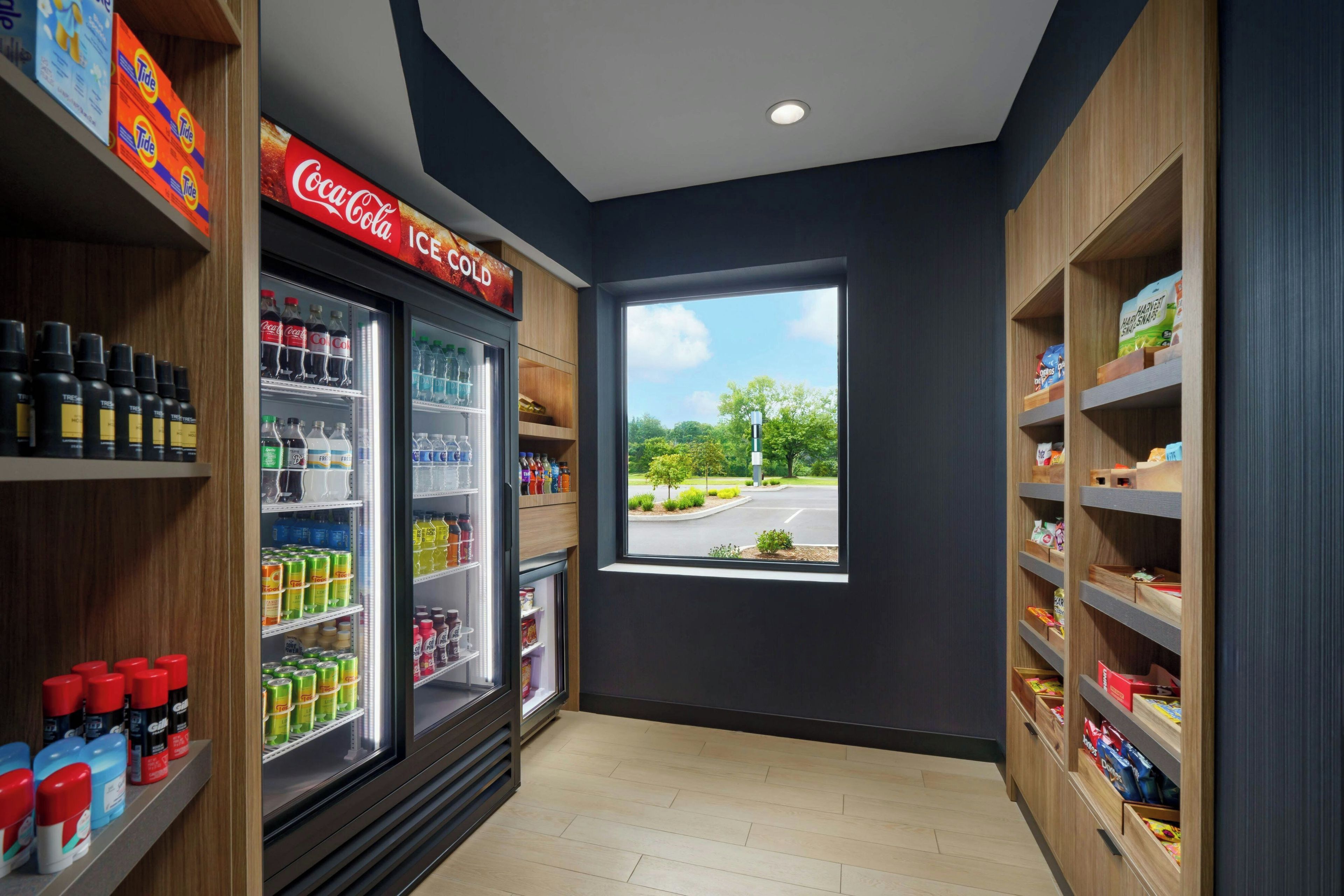 L'intérieur d'un magasin, avec des étagères réfrigérées remplies de boissons et d'en-cas, un congélateur Coca-Cola, et une vue sur un parking à travers une fenêtre.
