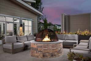 A stone fire pit surrounded by grey sofas and chairs, with a view of a modern house and a pink sky in the background. - Residence Inn By Marriott Ocean Township (Oakhurst)