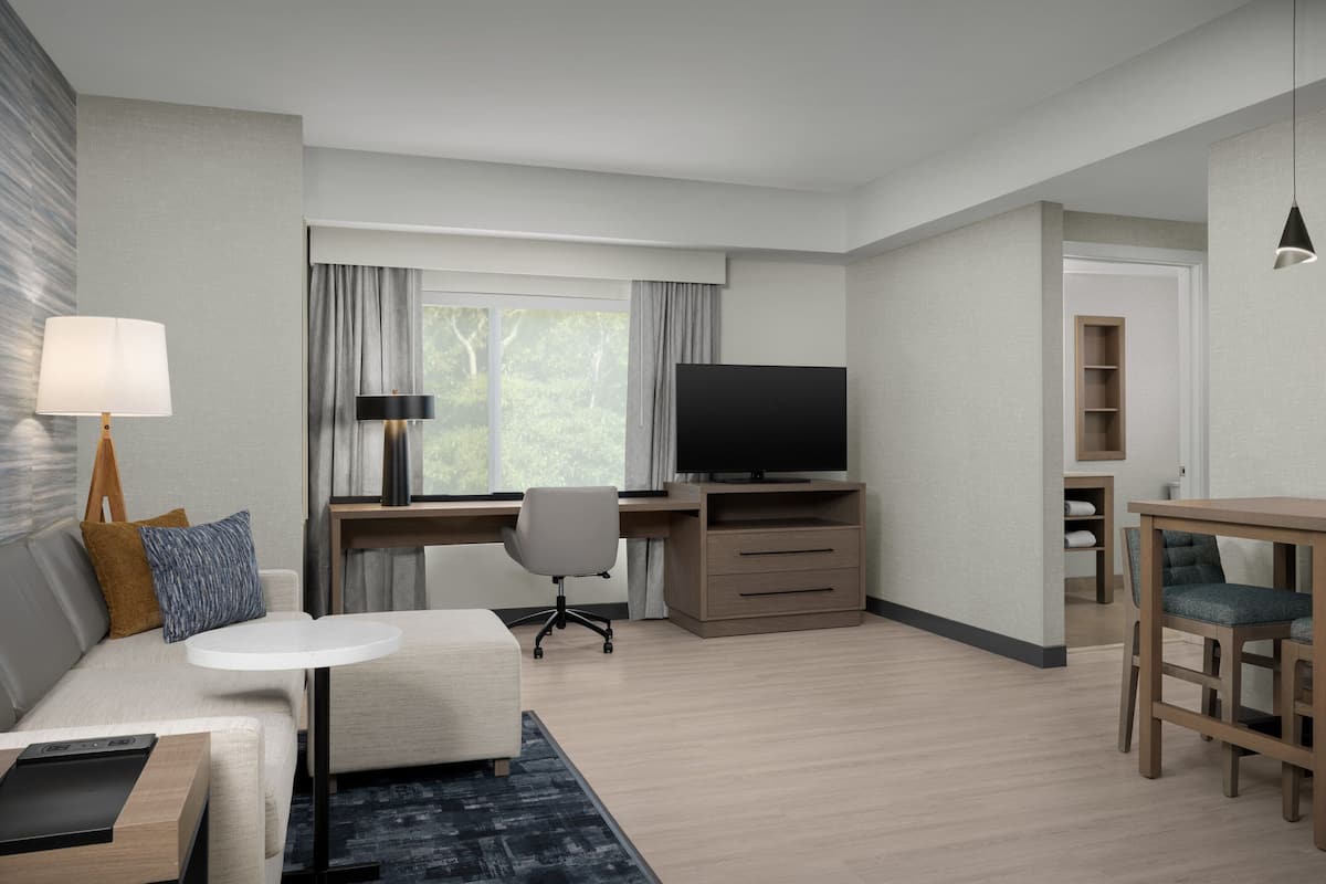 Quarto de hotel moderno com sofá, uma pequena mesa, escrivaninha com computador, televisão e estante de livros.