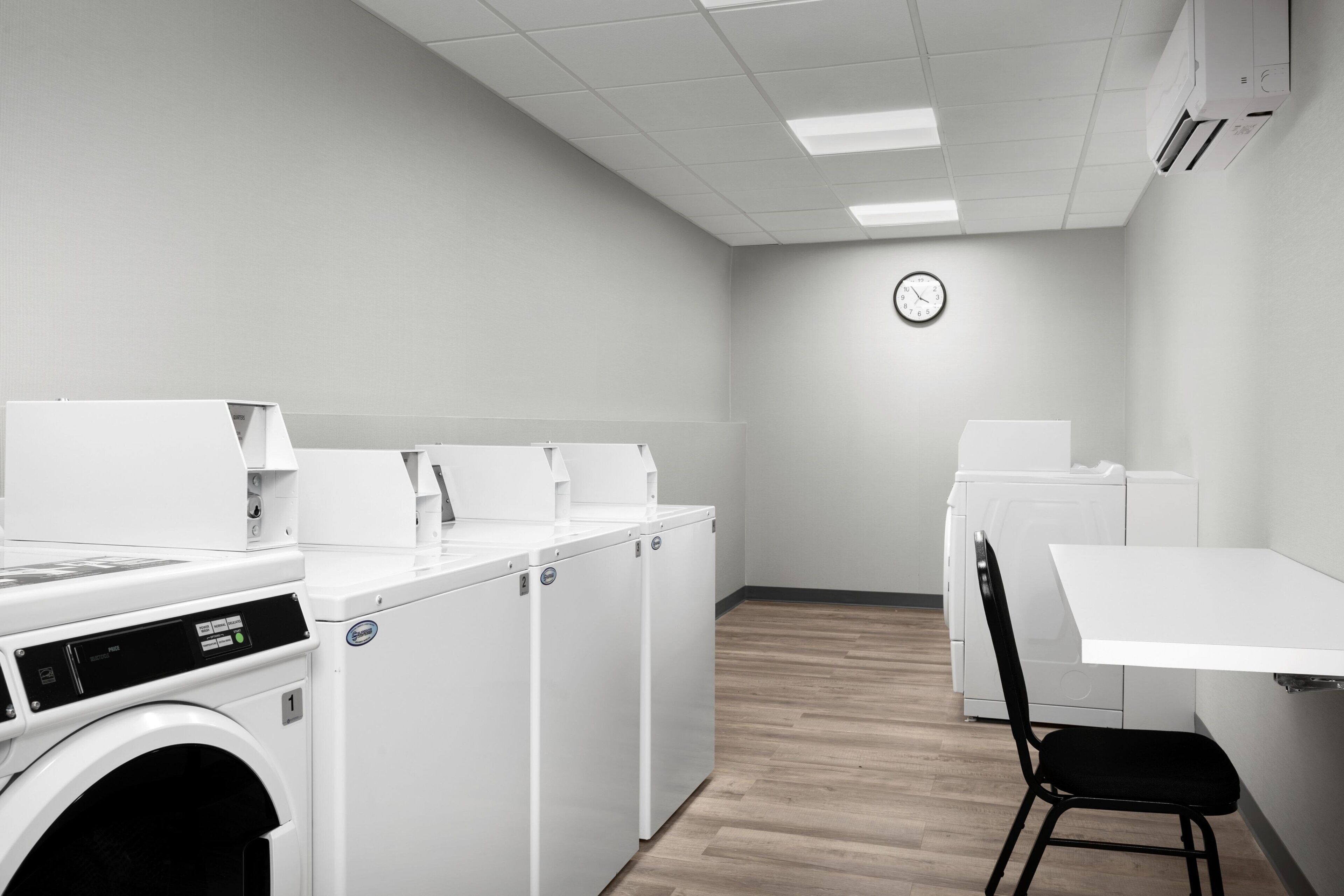 A laundry room with multiple washing machines and dryers, a clock on the wall, and a table with a chair.