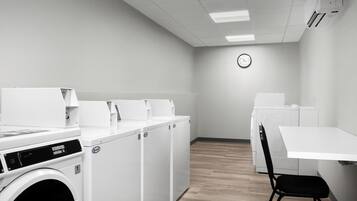 A laundry room with multiple washing machines and dryers, a clock on the wall, and a table with a chair.