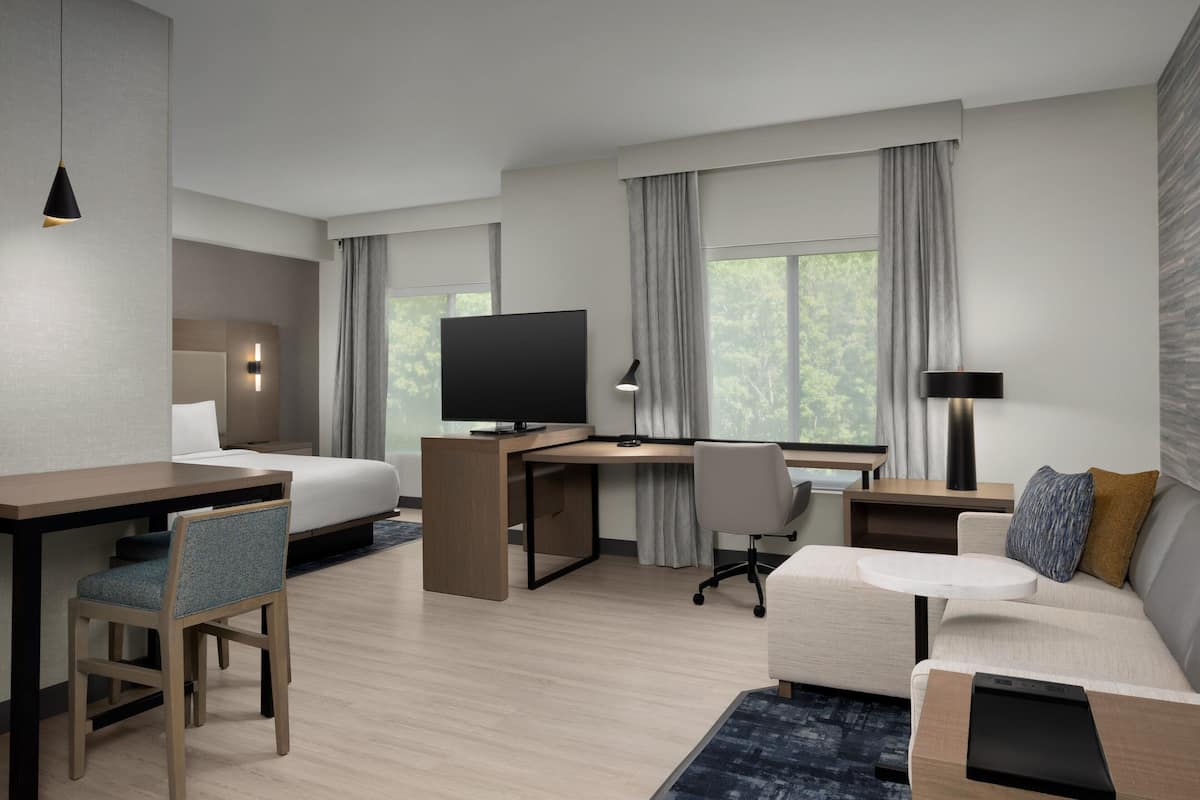 Quarto de hotel moderno com cama, escrivaninha, cadeira, TV e sofá.