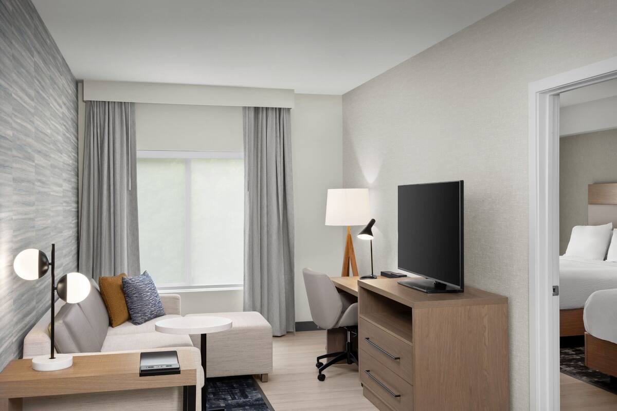 Quarto de hotel moderno com sofá, escrivaninha, televisão, luminária e cama.