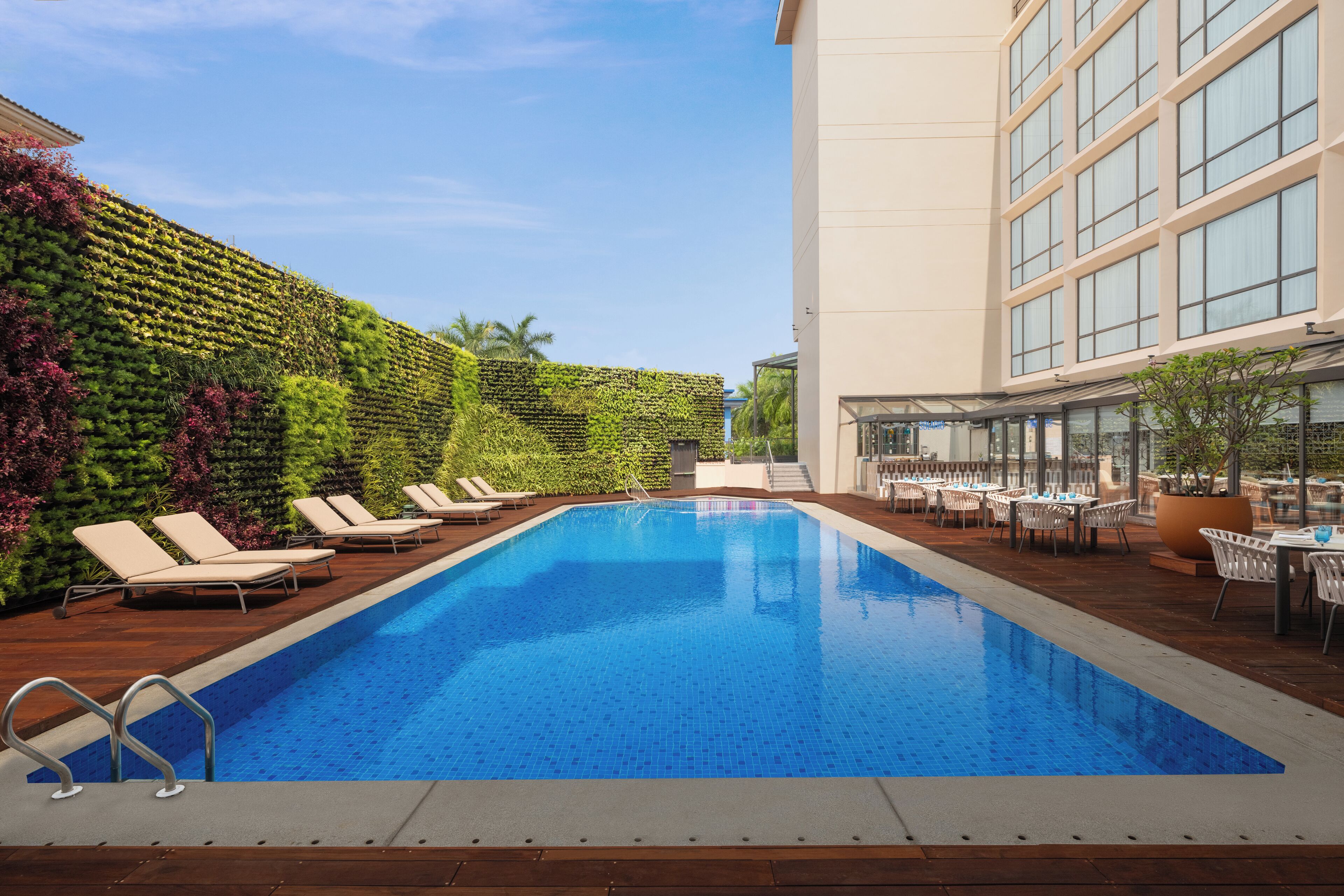 A swimming pool with lounge chairs and tables, surrounded by a building with large windows and a green wall.