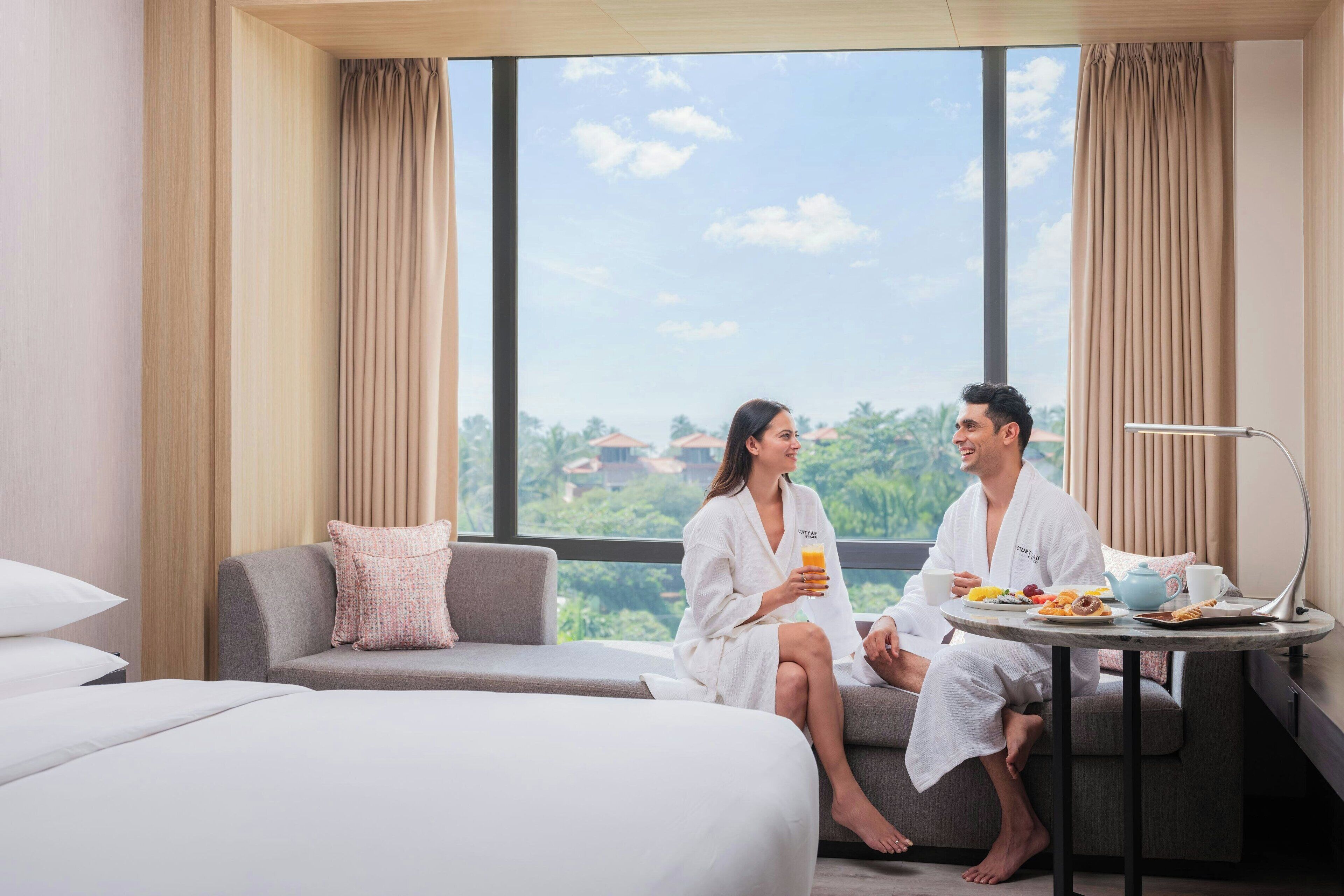 A hotel room with a large window, a sofa, a small round table with breakfast items, and a bed with white linens.
