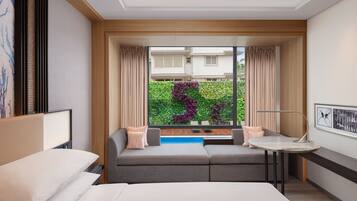 Habitación de hotel moderna con una cama, un sofá, una mesita y vistas a un jardín verde a través de un ventanal amplio.