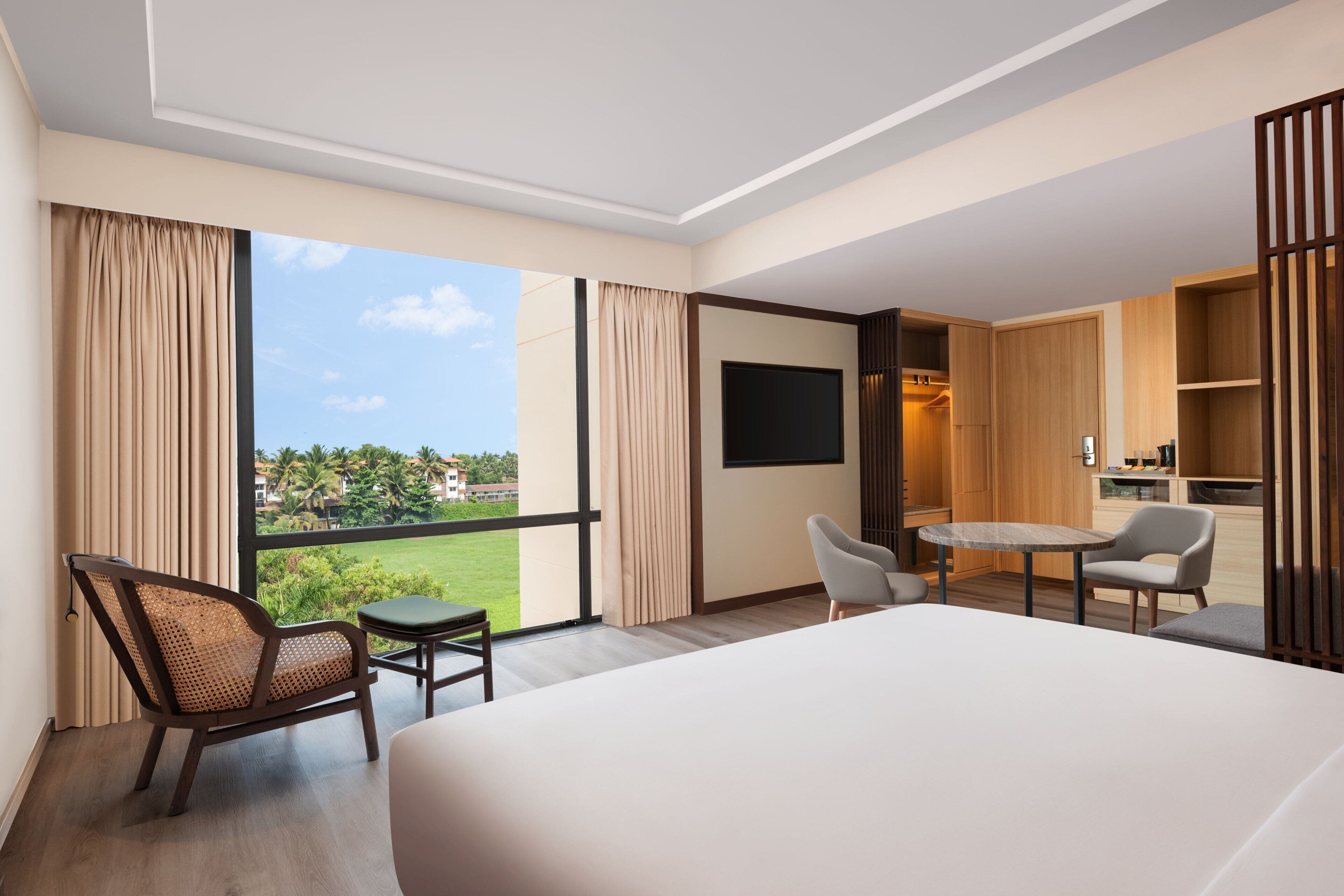 A hotel room with a large bed, a dining table, two chairs, a TV, and a view of a green landscape.