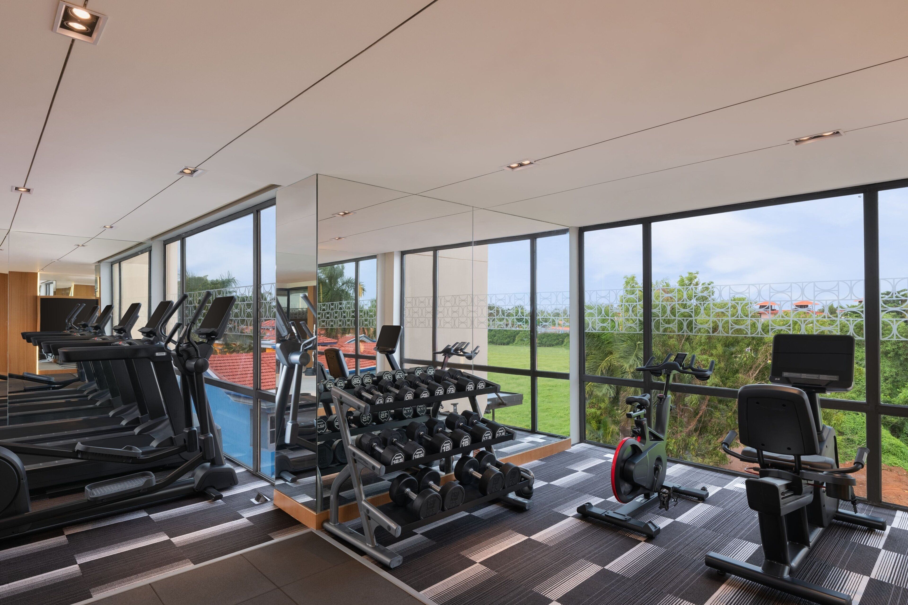 A gym with treadmills, dumbbells, and exercise equipment, large windows, and a view of greenery.