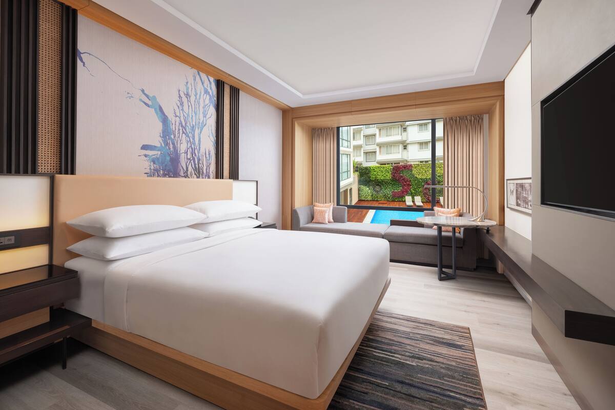 A modern hotel room with a large bed, a small table, a sofa, a TV, and a window with a view of other buildings and greenery.