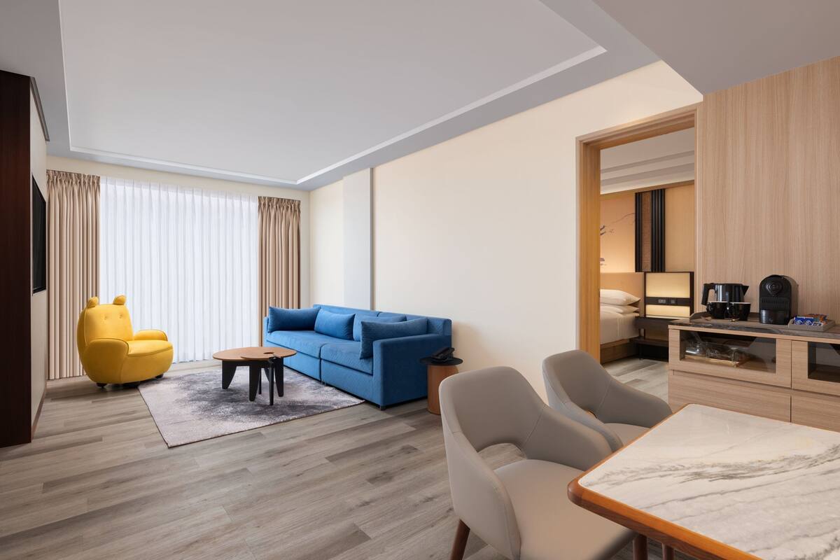 A modern hotel room with a blue sofa, a yellow chair, a marble table, and a kitchenette area.