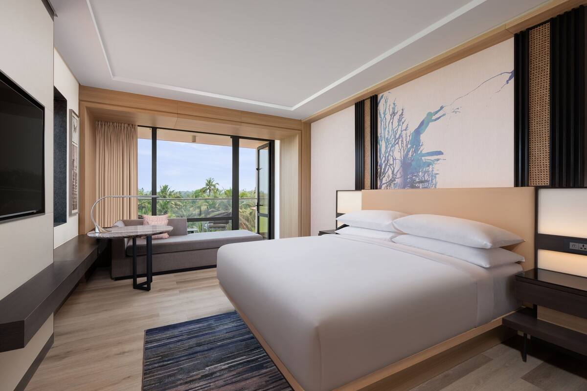 A modern hotel room with a large bed, a desk, and a window offering a view of greenery.