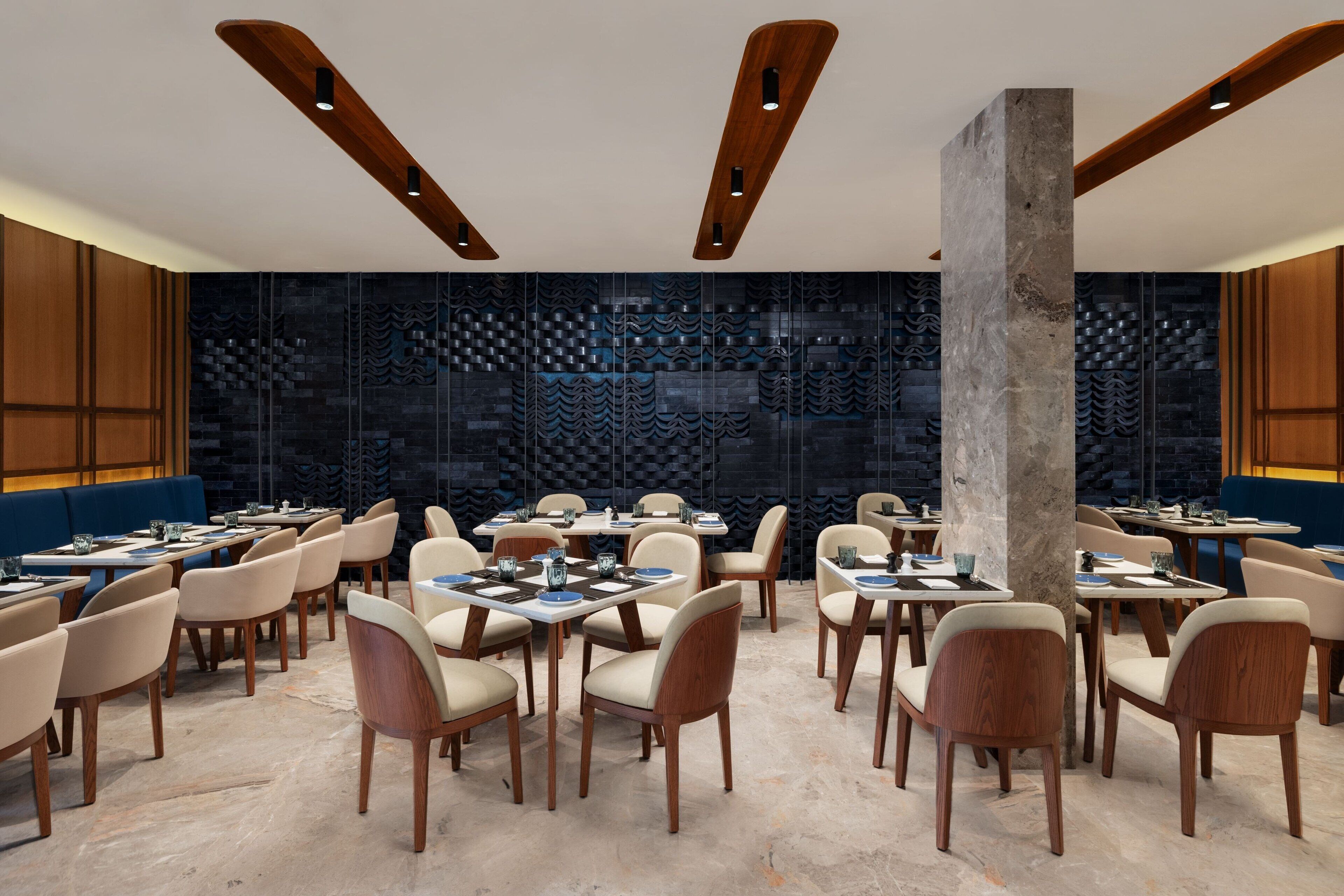 Uma sala de jantar moderna com vigas de madeira no teto, mesas dispostas com louças azuis e brancas, e cadeiras bege.