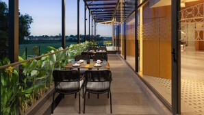An outdoor dining area with tables and chairs, a glass railing, and a view of the landscape. - Courtyard By Marriott Goa Colva (Colva)