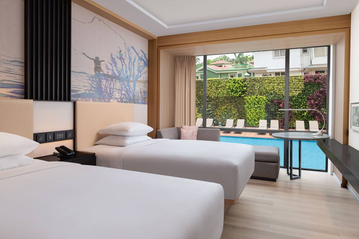 A hotel room with two beds, a small table, and a view of a swimming pool and greenery outside.