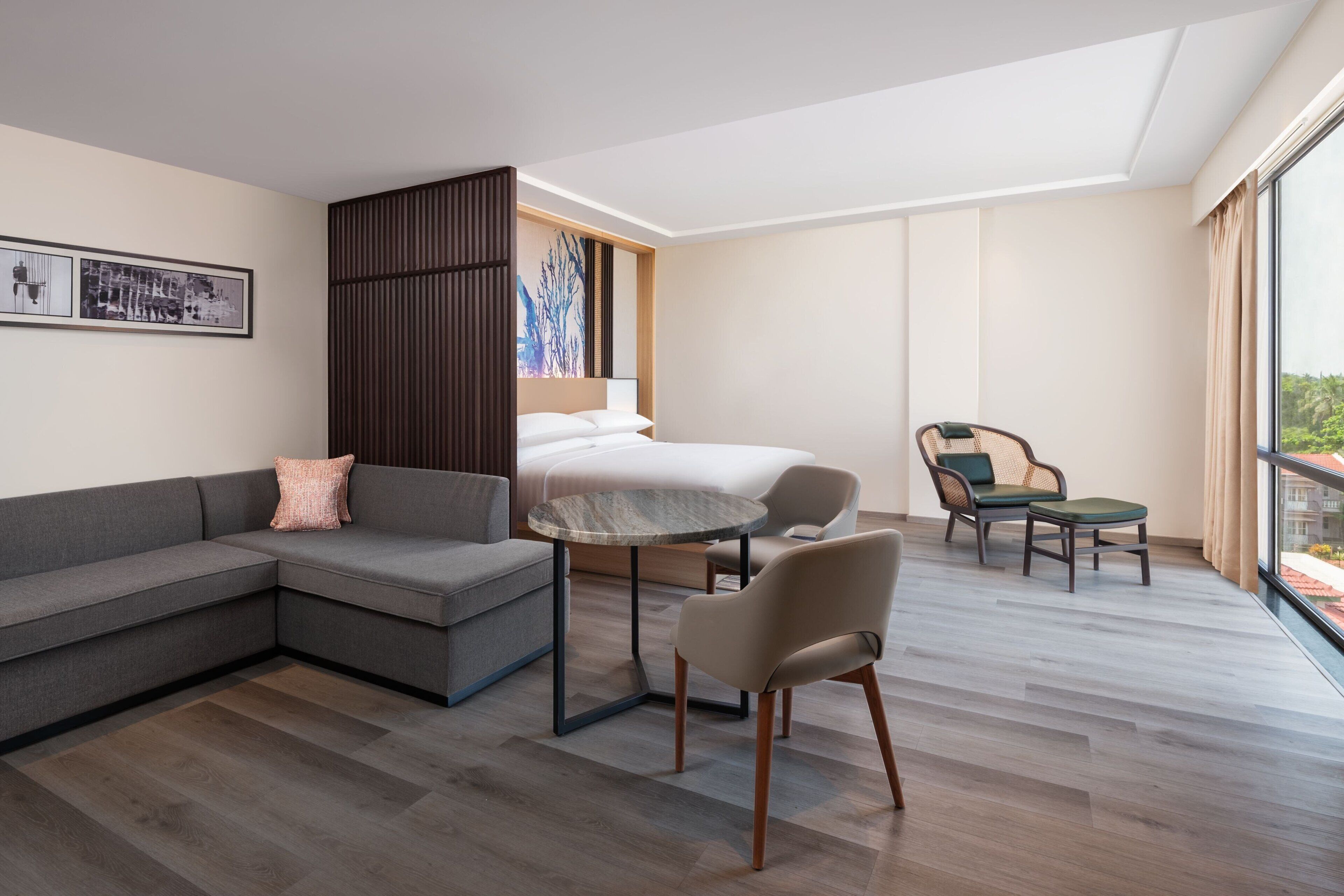 A modern hotel room with a sectional sofa, a round table, a chair, a bed, and a large window offering a view.