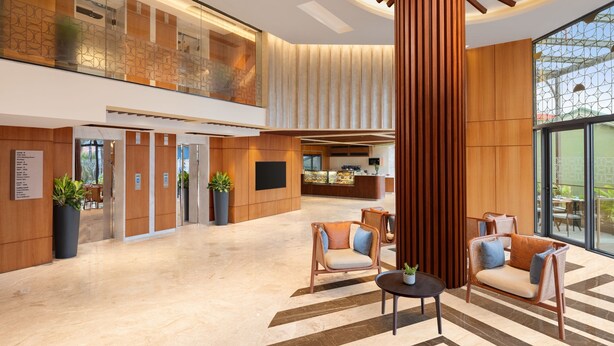 A modern lobby with a high ceiling, wooden paneling, a central column, seating area, and a reception desk. - Courtyard By Marriott Goa Colva (Colva)