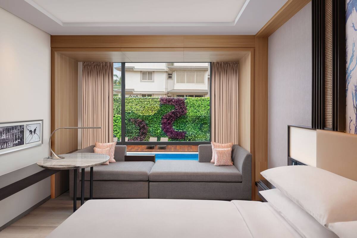 Et moderne hotelværelse med sofa, lille bord, seng og udsigt til en have gennem et stort vindue.