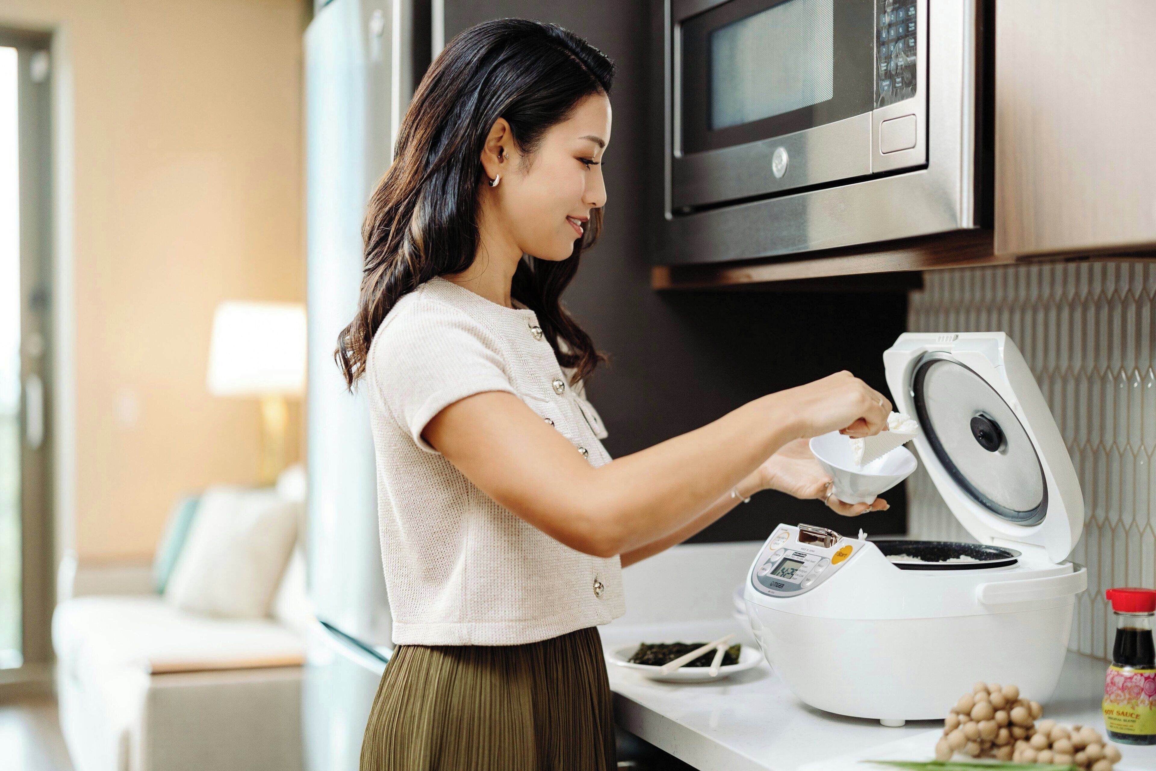 Uma pessoa cozinha em uma cozinha moderna, com um cozedor de arroz, micro-ondas e uma mesa de jantar com um prato de comida e um frasco de molho.