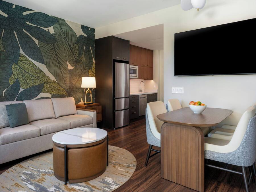 Una sala de estar moderna con un sofá, una mesa de centro y un comedor con sillas. Se ve una cocina con un refrigerador y un microondas al fondo.