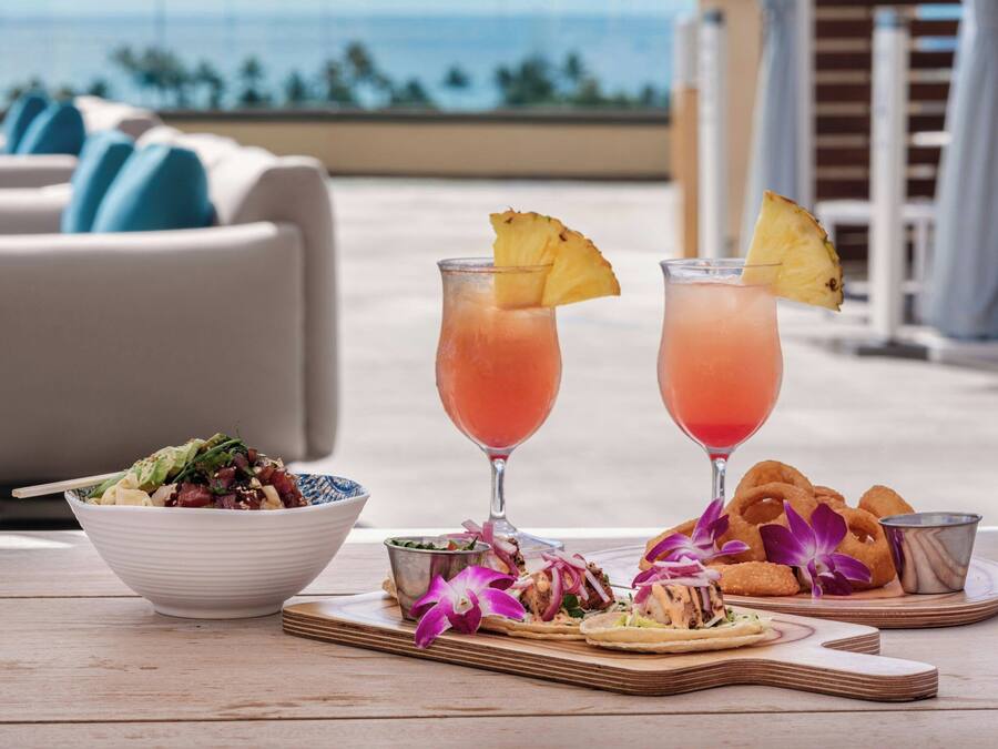 Een tafel met een saladeschaal, twee drankjes versierd met ananas, en een bord chips met een klein bakje salsa.