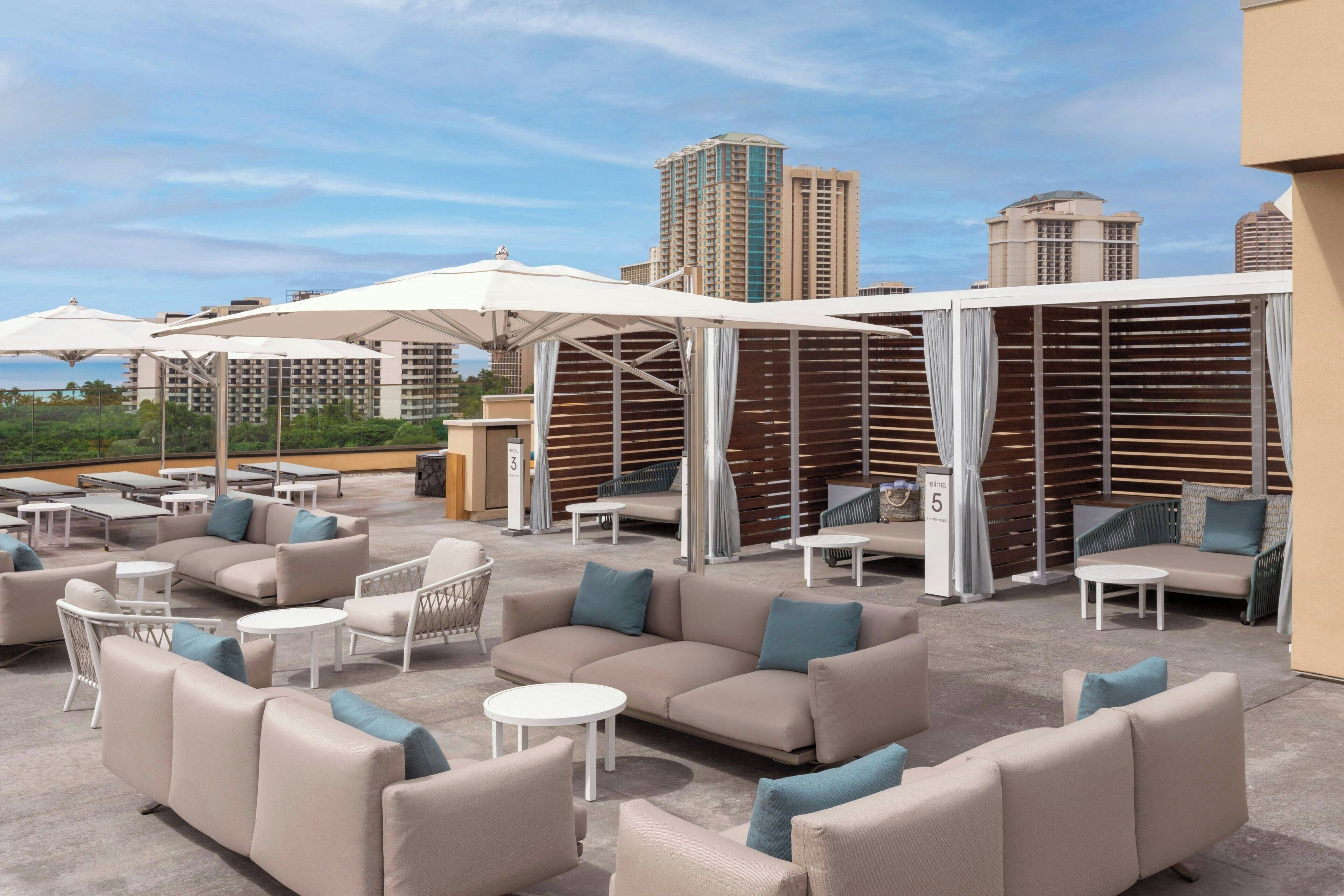 Un espacio lounge al aire libre con diversas opciones de asientos, incluyendo sofás y sillones, dispuestos alrededor de mesas y sombrillas.
