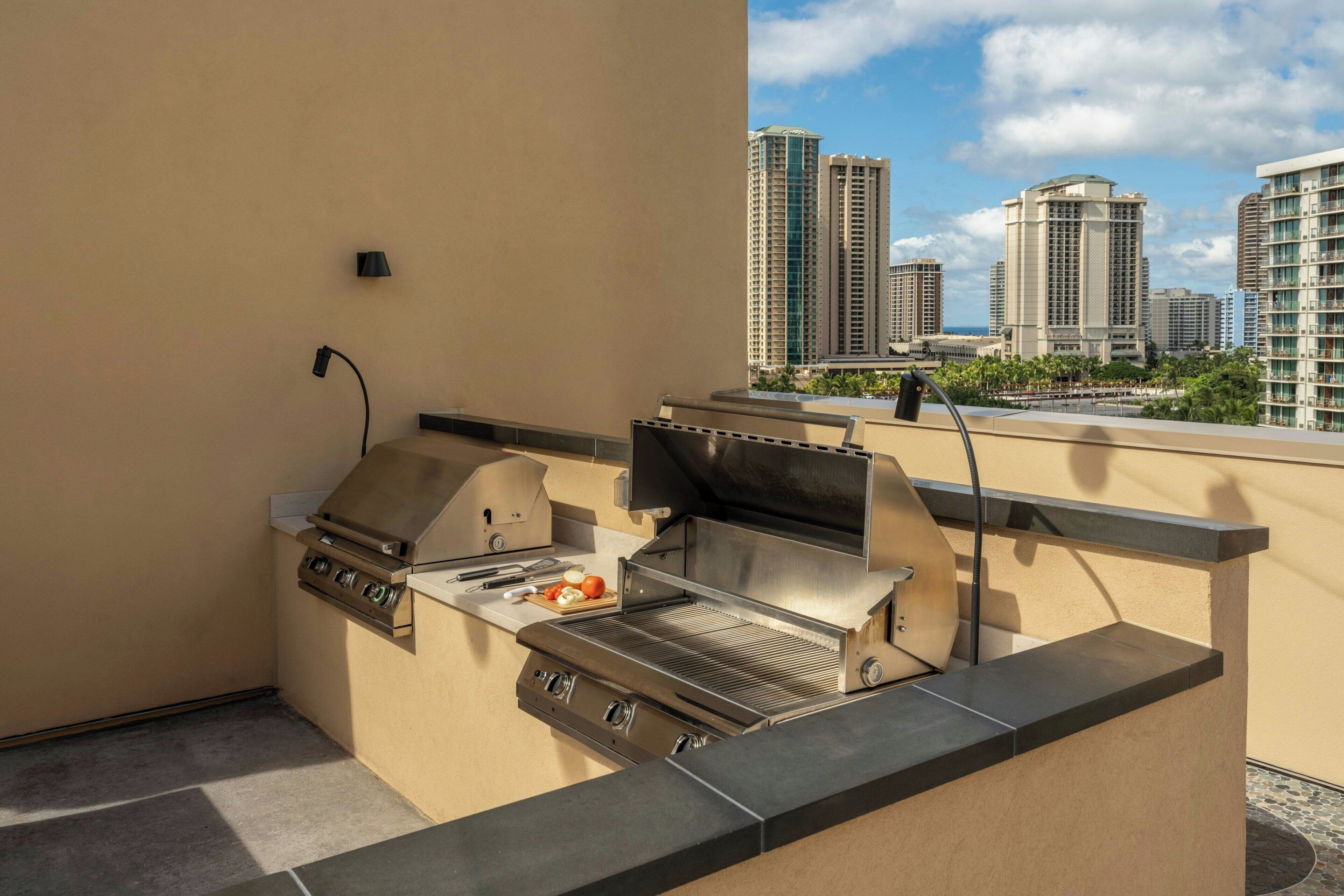 An outdoor kitchen with a grill and a sink, overlooking a cityscape with tall buildings.