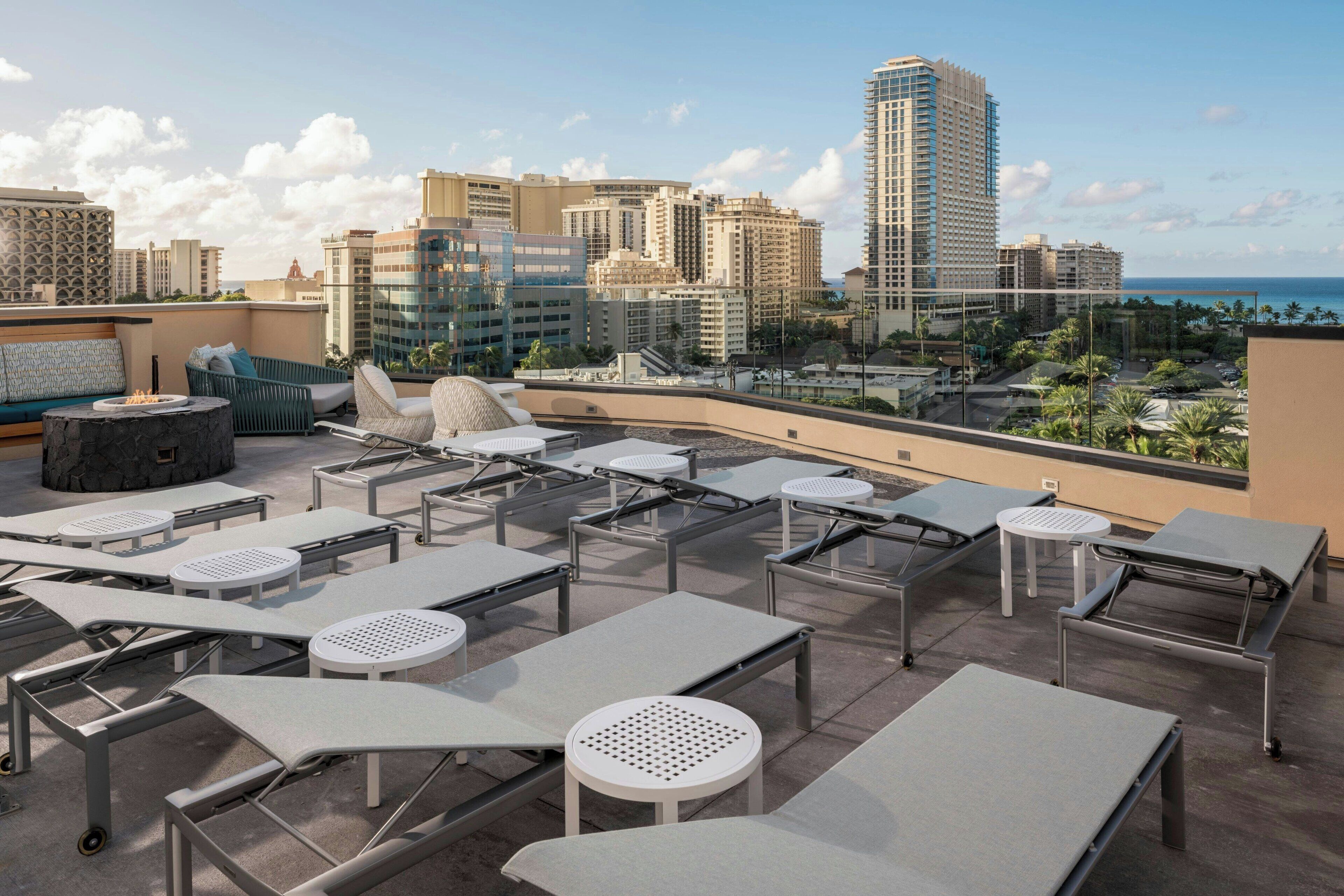 A rooftop lounge area with sun loungers, tables, and chairs, overlooking a cityscape and the ocean.