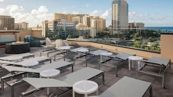 A rooftop lounge area with sun loungers, tables, and chairs, overlooking a cityscape and the ocean.