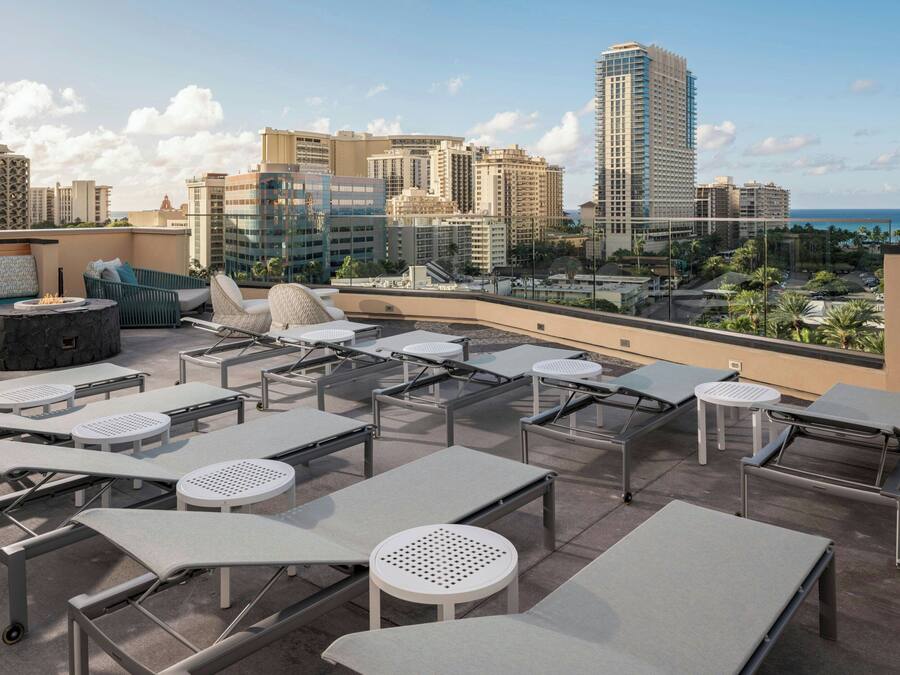 A rooftop lounge area with sun loungers, tables, and chairs, overlooking a cityscape and the ocean.