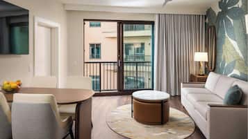 Habitación de hotel con comedor, sofá, otomana redonda, ventilador de techo, balcón con vistas a otros edificios y un ventanal con cortinas.