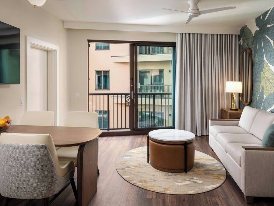 Habitación de hotel con comedor, sofá, otomana redonda, ventilador de techo, balcón con vistas a otros edificios y un ventanal con cortinas.