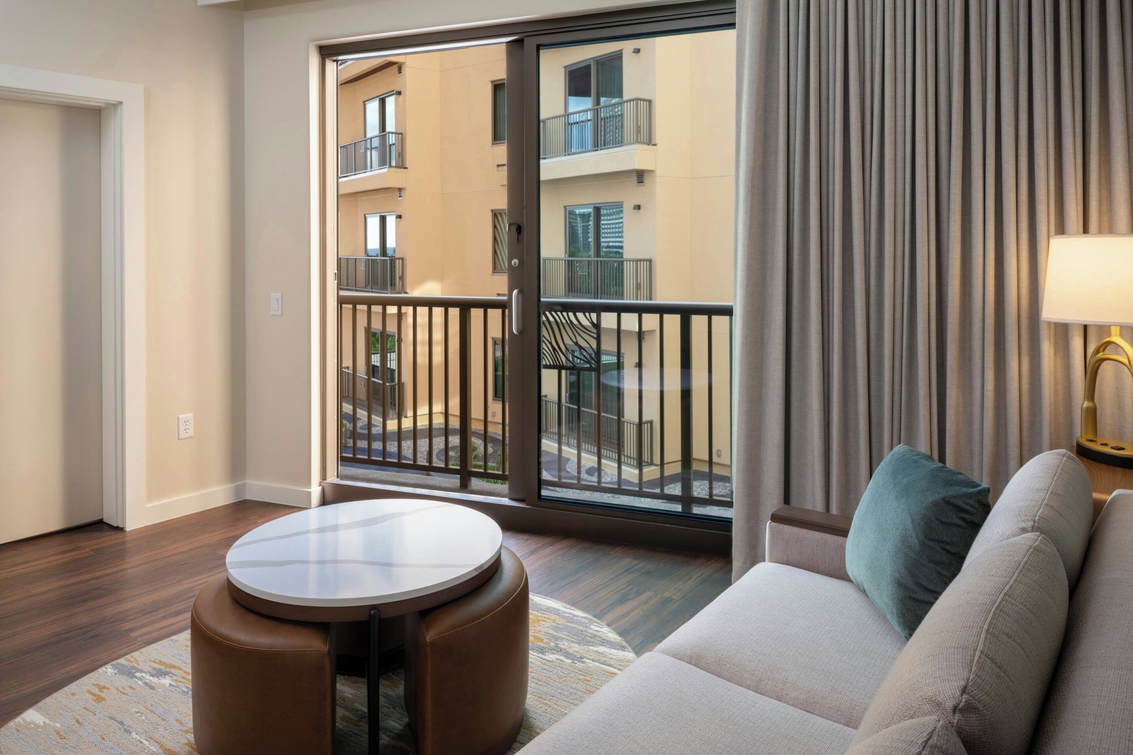 A modern hotel room with a large sliding glass door to a balcony, a round table, a sofa, and a lamp.