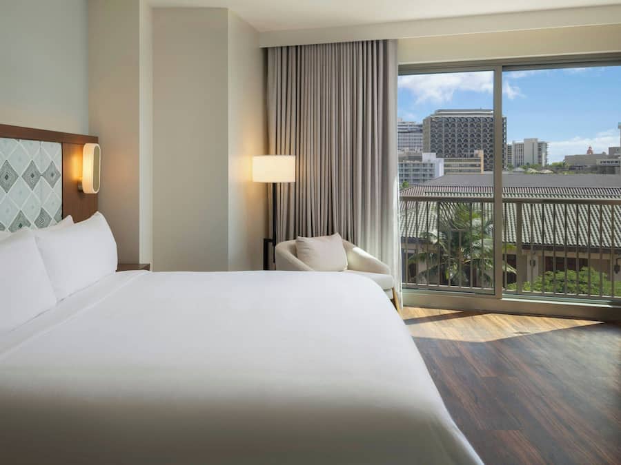 A hotel room with a large bed, a chair, a view of the city through a sliding glass door, and a patterned wall.