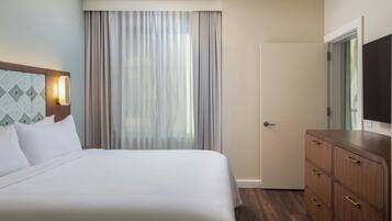 A hotel room with a bed, a wooden dresser, a door, and a window with curtains.