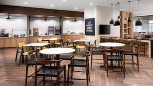 Un restaurante moderno con suelo de madera, mesas redondas y sillas. Hay lámparas colgantes y una pizarra con una frase motivacional.