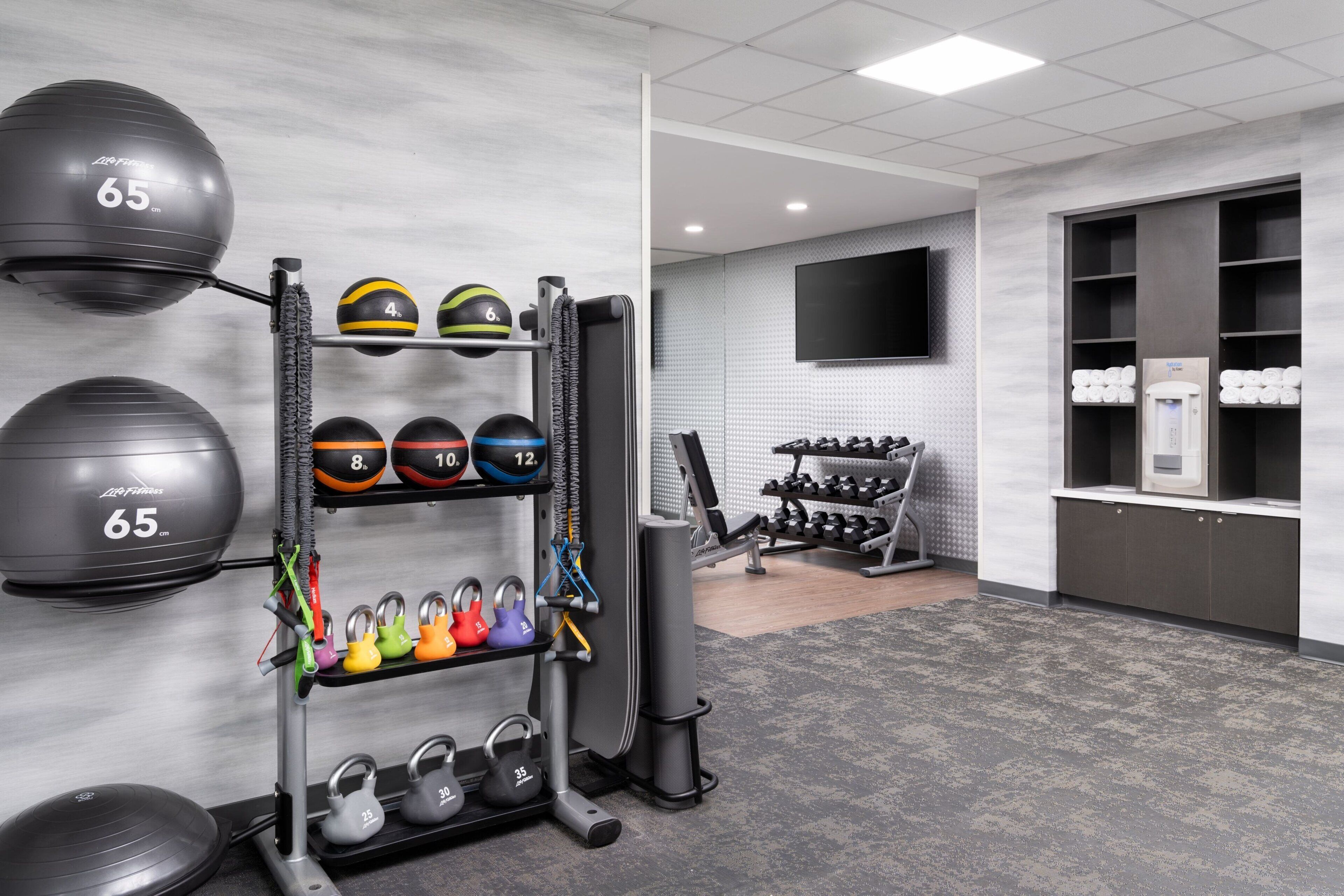 A gym with exercise equipment, including a rack of dumbbells and medicine balls, and a TV mounted on the wall.