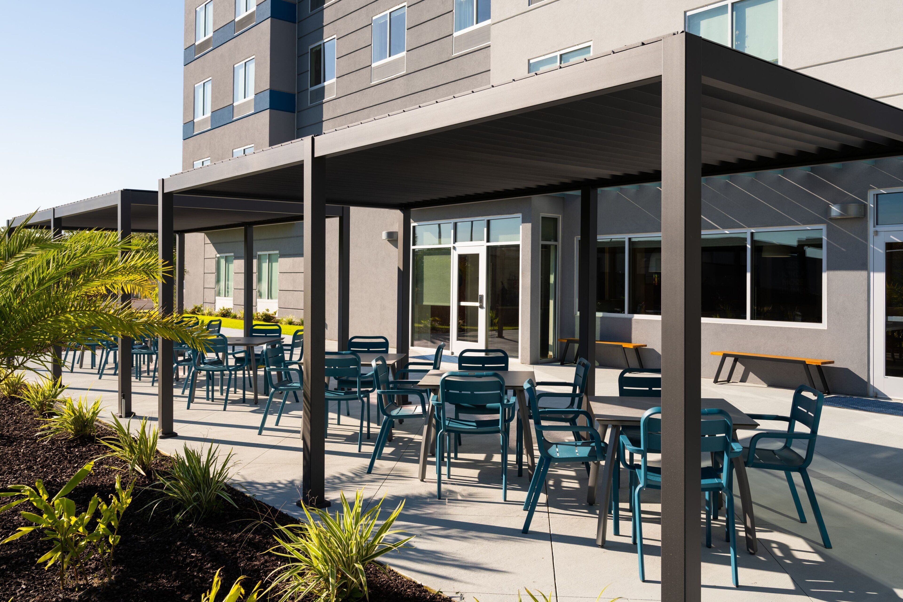 An outdoor patio area with tables and chairs, surrounded by a building with large windows and a covered walkway.