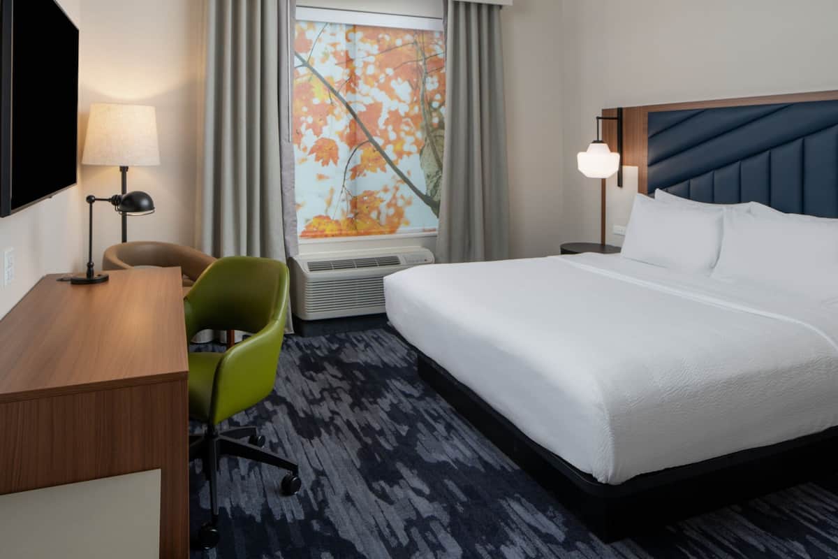 A hotel room with a bed, a desk, a chair, a TV, a window with curtains, and a nightstand with a lamp.