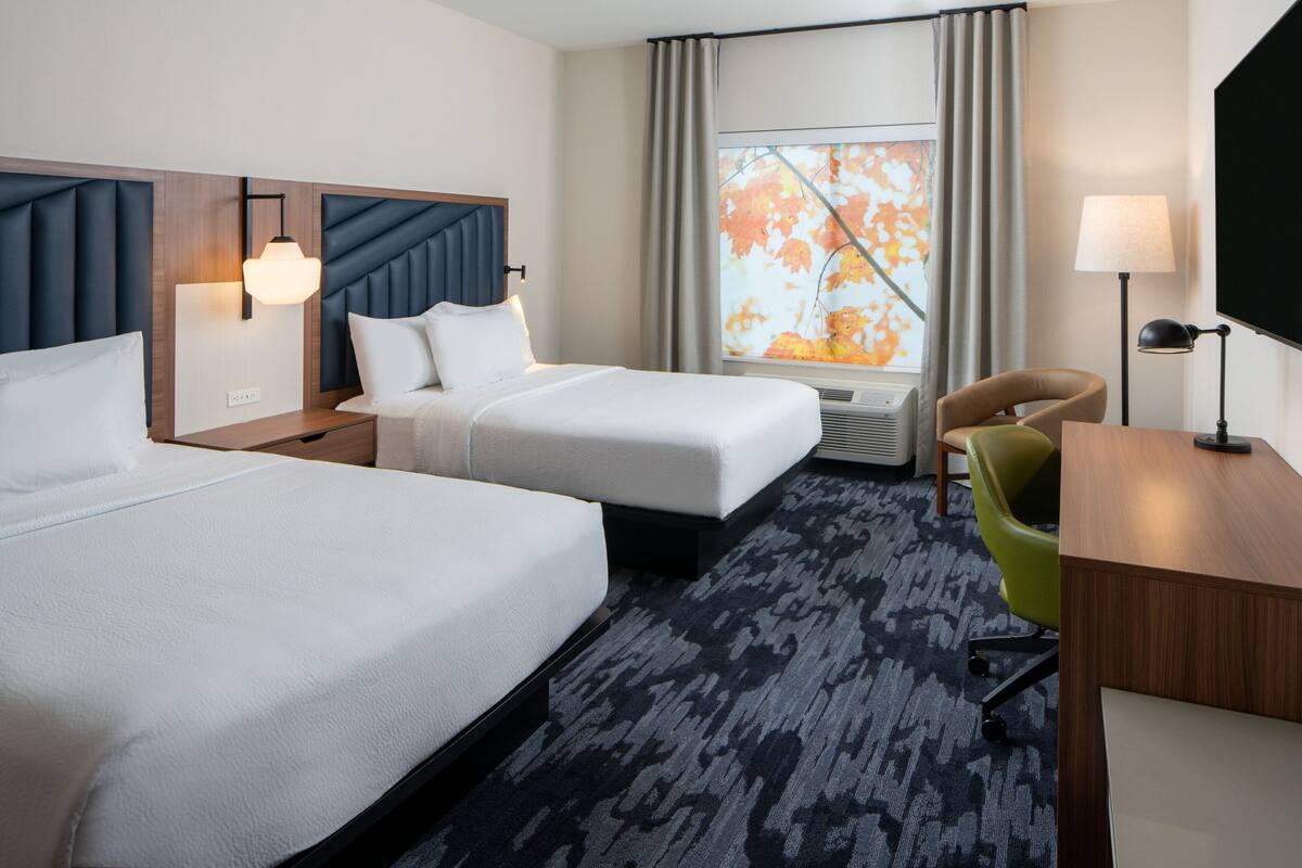 A hotel room with two beds, a desk, a chair, a TV, and a window with a view of autumn leaves.