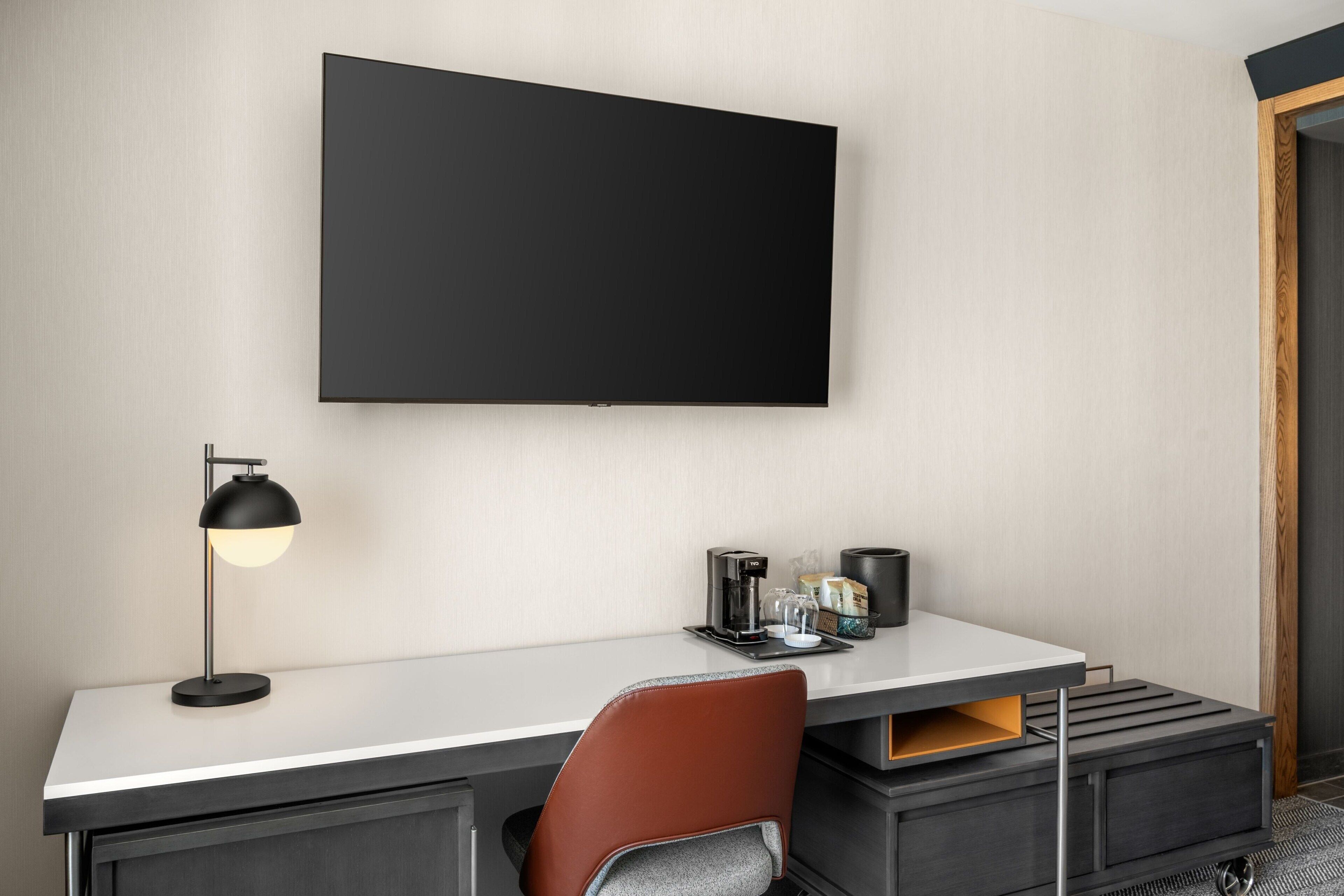 A modern hotel room with a flat-screen TV mounted on the wall, a desk with a lamp, a chair, and a coffee maker.