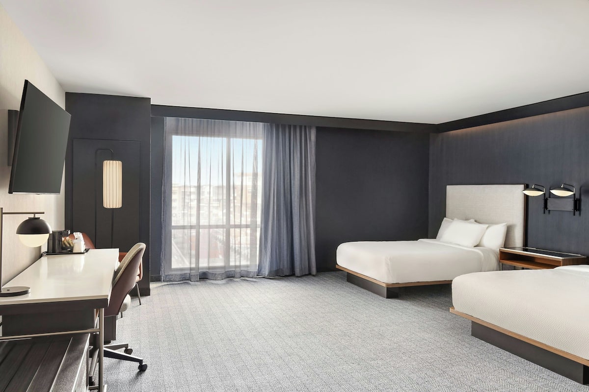 Een hotelkamer met een bureau, twee bedden, een groot raam met gordijnen en uitzicht op de stad.