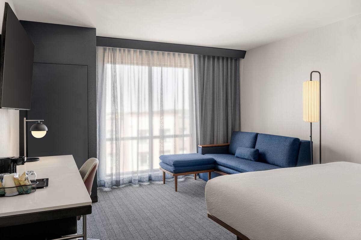 Hotelkamer met een bed, bureau, stoel, bank, lamp en een groot raam met gordijnen.