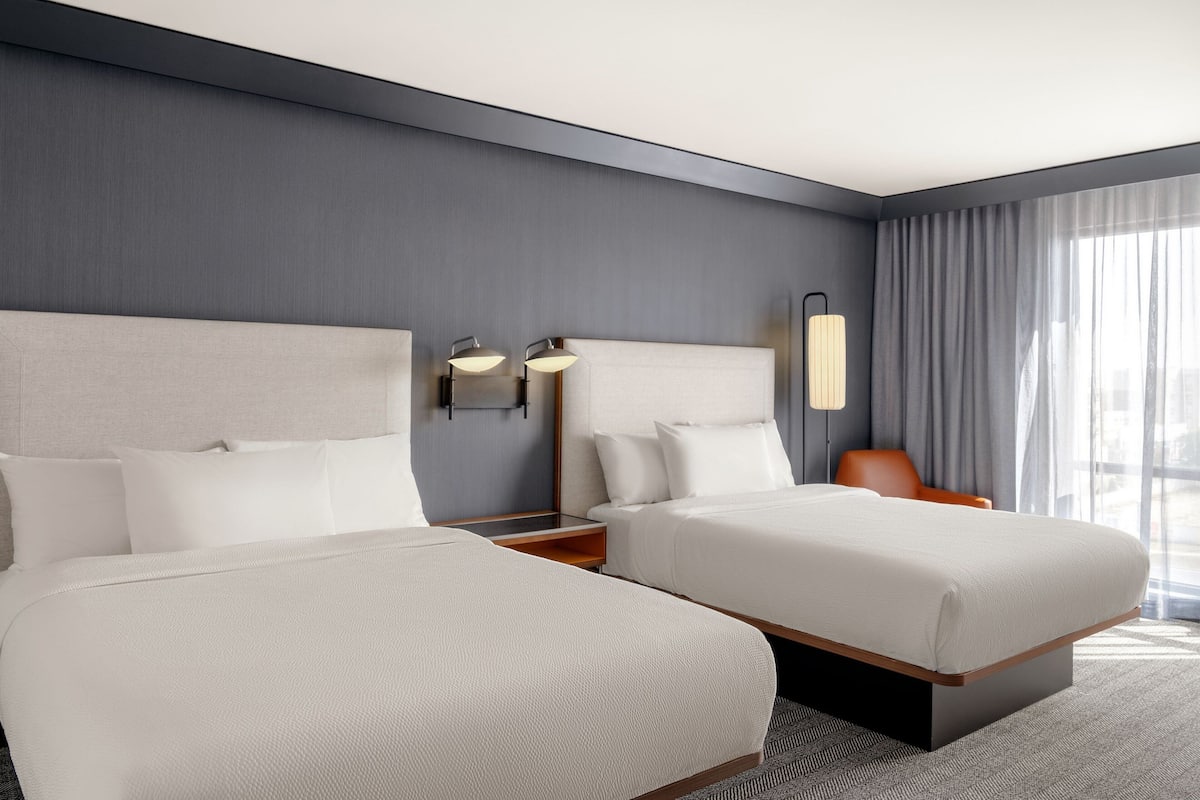 Een moderne hotelkamer met twee bedden, een nachtkastje, een lamp en een groot raam met gordijnen.
