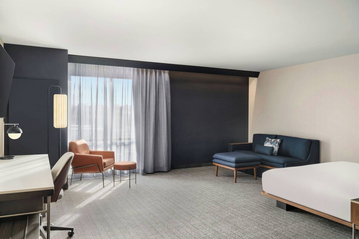 Een hotelkamer met een bed, een bureau, een stoel, een bank en een groot raam met gordijnen.
