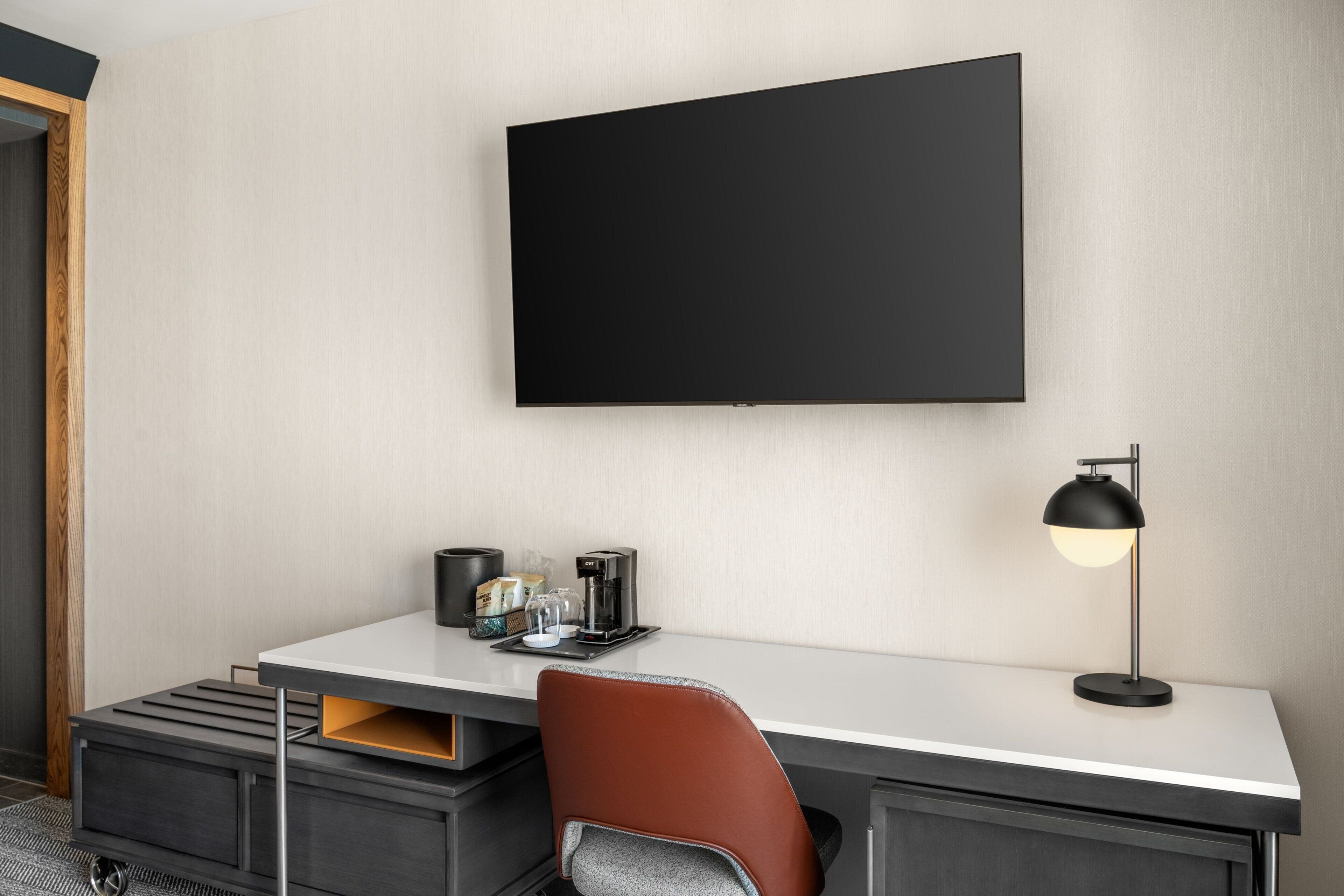 Una habitación de hotel moderna con una televisión de pantalla plana montada en la pared, un escritorio con cafetera, una lámpara y un sillón de cuero marrón.