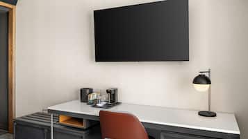 Una habitación de hotel moderna con una televisión de pantalla plana montada en la pared, un escritorio con cafetera, una lámpara y un sillón de cuero marrón.
