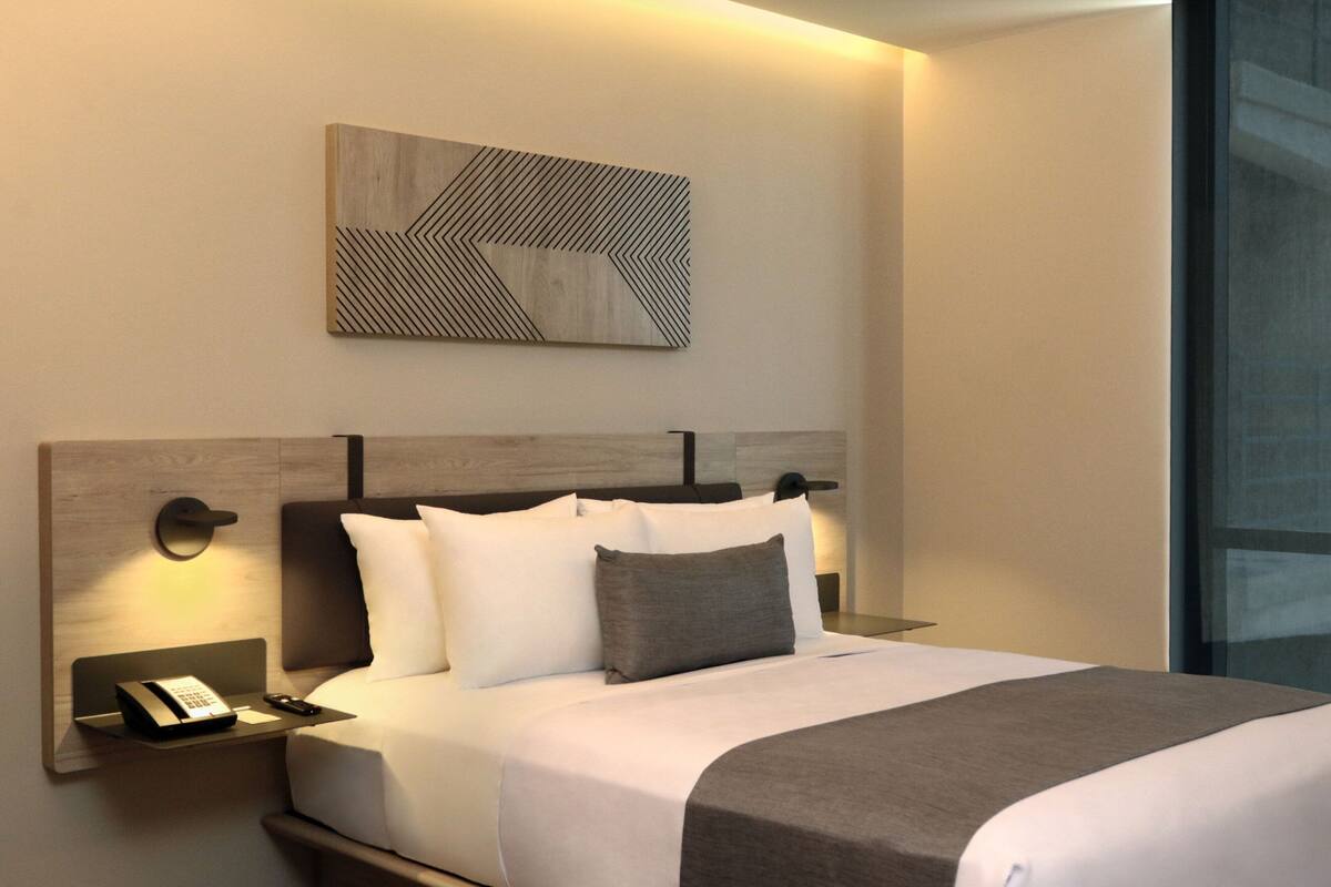 A modern hotel room with a neatly made bed, a bedside table with a phone and remote, and a wall-mounted artwork.