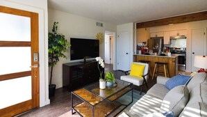 A modern living room with a sofa, a coffee table, and a TV. There is a kitchen area with a bar and a dining table.