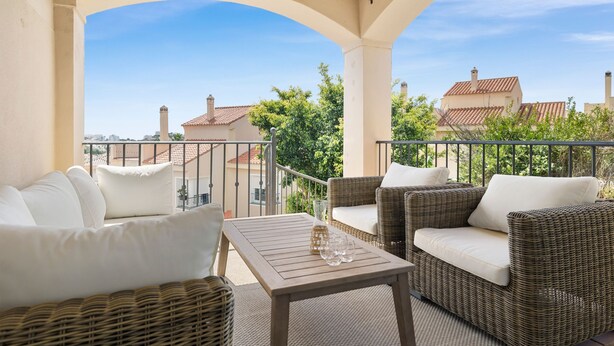 Un balcón con mobiliario de mimbre y una mesa, con vistas a una zona residencial con casas y árboles.